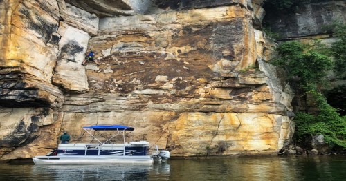 A person climbs a rocky cliff beside a boat on calm water, surrounded by lush greenery and steep rock formations.