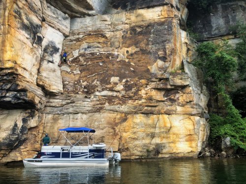 A boat anchored near a rocky cliff, with a person climbing the vertical rock face above the water.