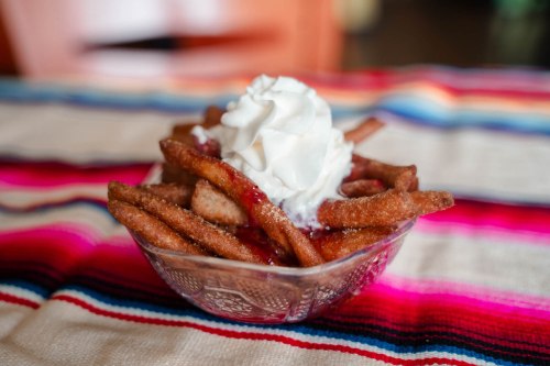 A bowl of fried churros topped with whipped cream and strawberry sauce, placed on a colorful striped tablecloth.