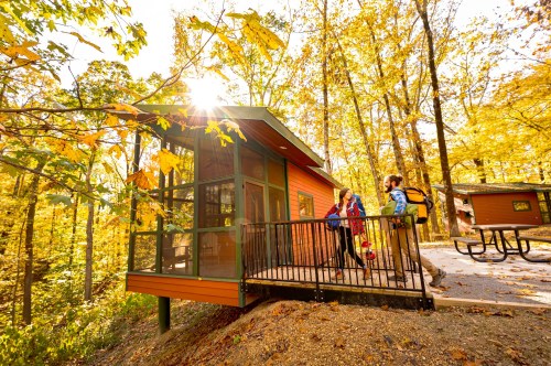 A modern cabin surrounded by autumn trees, with three people chatting on the deck in warm sunlight.