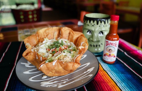 A colorful plate featuring a taco salad in a crispy shell, topped with lettuce, tomatoes, and a bottle of hot sauce.