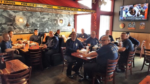 A group of firefighters sitting together at a restaurant, enjoying a meal and watching a game on TV.