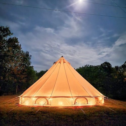 A lit tent stands in a grassy area under a night sky, with trees in the background and a bright moon above.