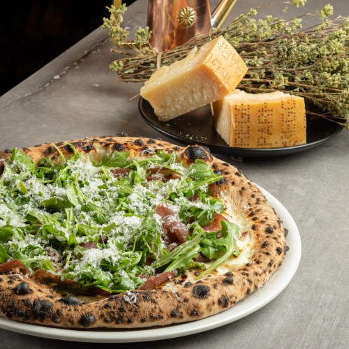 A pizza topped with fresh greens, cheese, and prosciutto, served on a white plate with cheese and herbs in the background.