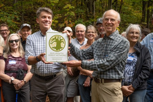 Two men hold a plaque for the Old-Growth Forest Network, surrounded by a group of smiling people in a forest setting.