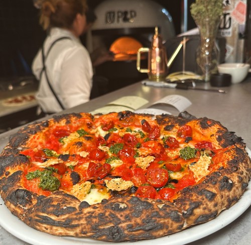 A freshly baked pizza with toppings, including cherry tomatoes and herbs, in front of a wood-fired oven.