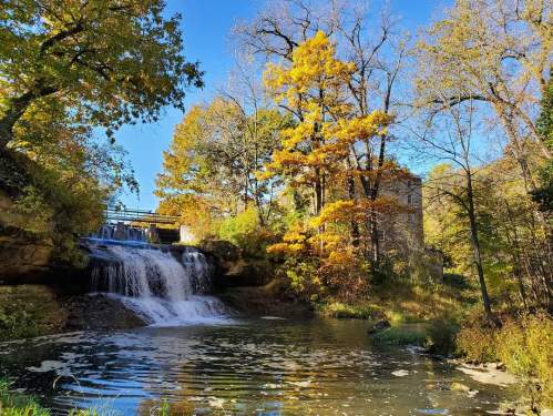A serene waterfall cascades into a pond, surrounded by vibrant autumn trees and a clear blue sky.