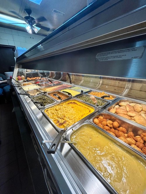 A buffet display featuring various dishes, including macaroni and cheese, greens, and fried items in metal trays.