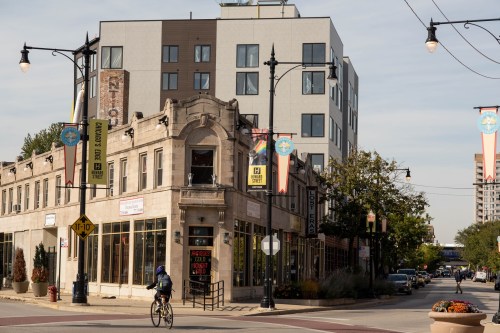 A cyclist rides past a historic building with modern apartments above, surrounded by trees and street banners.