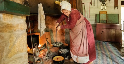 A woman in historical attire stirs a pot over a fireplace in a rustic kitchen, surrounded by cooking utensils.