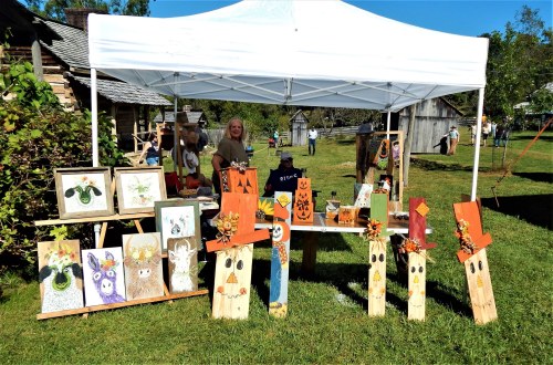 A craft booth under a white tent displays painted wooden decorations and artwork, with a vendor in the background.