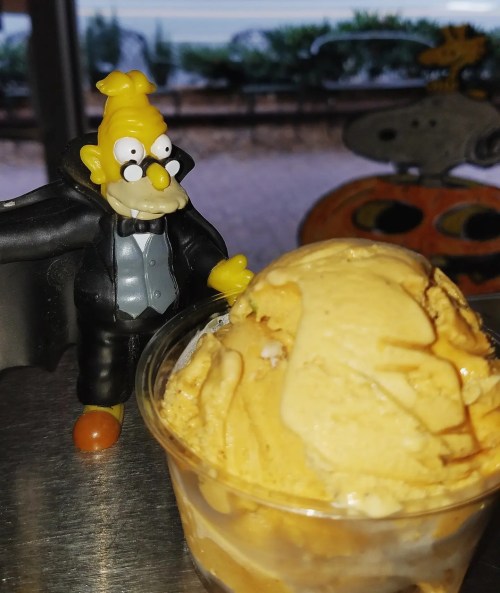 A toy figure of an elderly man in a tuxedo stands next to a cup of creamy caramel ice cream.