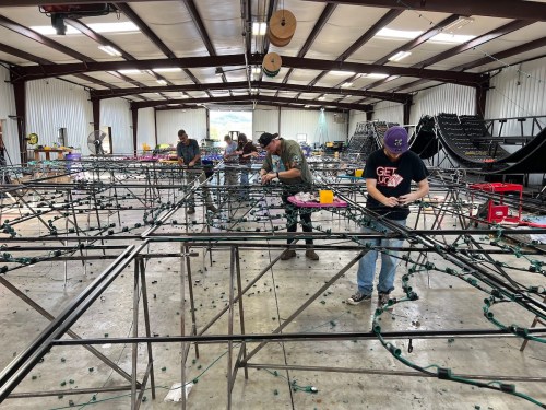 Workers assembling a large metal frame with lights in a spacious warehouse. Tools and materials are scattered around.