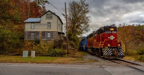 A red and black train passes by a rustic house surrounded by autumn foliage and a rural landscape.
