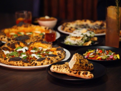 A table spread with various pizzas, salads, and flatbreads, showcasing a variety of toppings and colors.