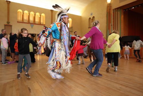A group of people, including a dancer in traditional attire, participate in a lively dance in a spacious hall.