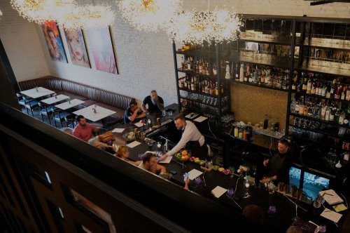 A bustling restaurant interior with a bar, patrons dining, and stylish chandeliers overhead.