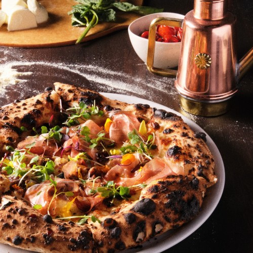 A freshly baked pizza topped with greens, meats, and colorful vegetables, set on a rustic wooden table.