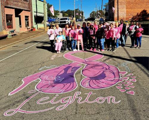 A group of people in pink attire stands on a street with a large pink ribbon mural, promoting breast cancer awareness.