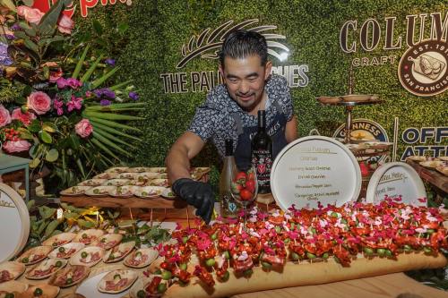 A chef arranges colorful appetizers on a table decorated with flowers and greenery at a food event.