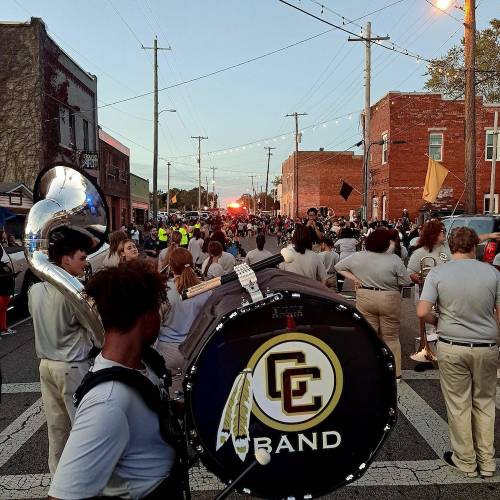 A marching band parades down a street at dusk, with musicians and spectators lining the road.