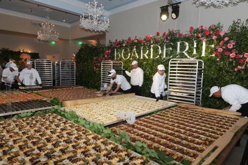 Caterers arrange an elaborate display of hors d'oeuvres at a lavish event, surrounded by floral decorations.