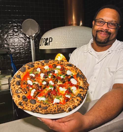A smiling chef holds a freshly baked pizza topped with cheese, herbs, and vegetables in a restaurant kitchen.