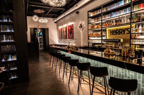A stylish bar with a sleek black countertop, colorful liquor shelves, and modern decor, featuring a cozy seating area.