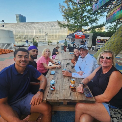 A group of six people sitting at a picnic table outdoors, enjoying drinks with a modern building in the background.