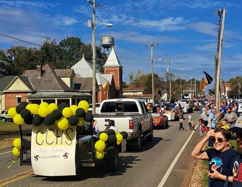 A parade scene with decorated vehicles and spectators, featuring a float with "CCHS Volleyball" and black and yellow balloons.