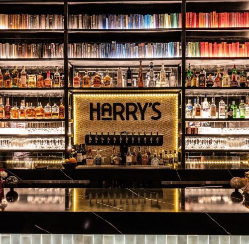 A stylish bar with a glowing sign reading "Harry's," surrounded by shelves of colorful liquor bottles.