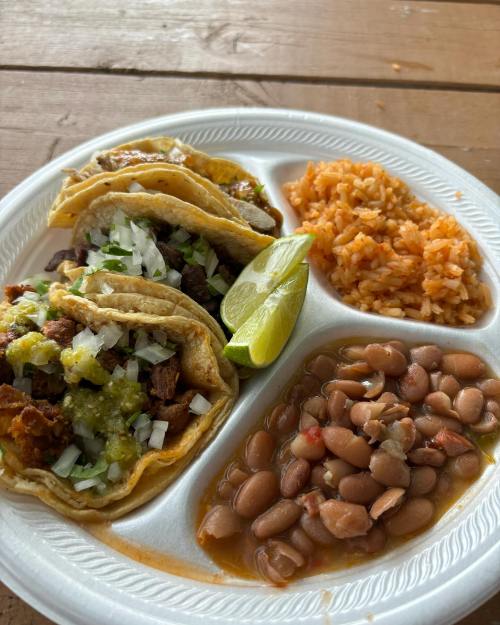 A plate with three tacos topped with onions and salsa, served with rice and pinto beans, garnished with a lime wedge.