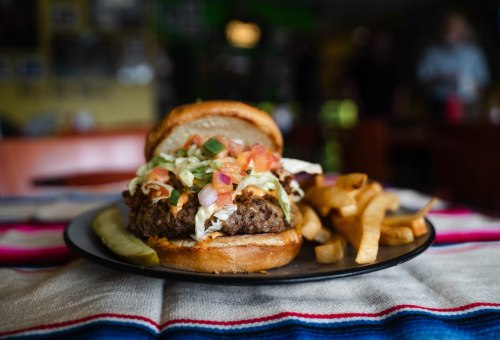 A close-up of a juicy burger topped with lettuce, tomato, and onions, served with fries and a pickle on a plate.
