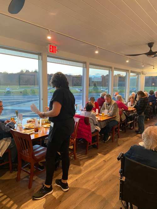 A busy restaurant interior with diners at tables and a server taking orders, large windows showing a scenic view outside.