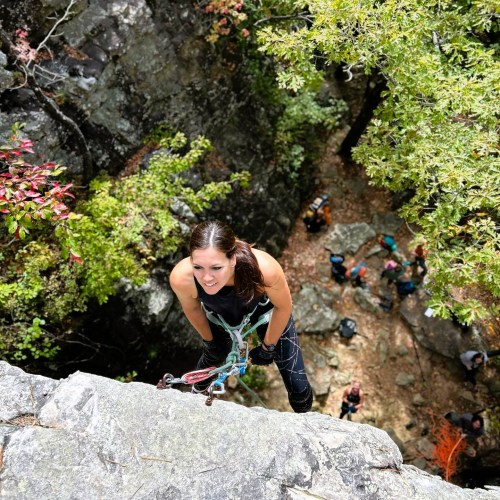 A climber ascends a rocky cliff, smiling, with trees and other climbers visible in the background.