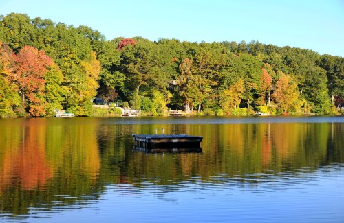 A serene lake reflects colorful autumn trees, with a small dock floating in the calm water.