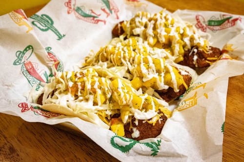 A basket of crispy fried food topped with cheese, sauce, and shredded lettuce, served on decorative paper.