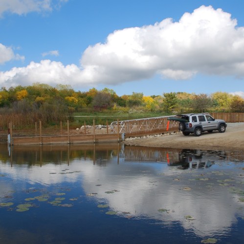 A serene landscape featuring a calm pond, a wooden dock, and a vehicle parked nearby under a blue sky with fluffy clouds.
