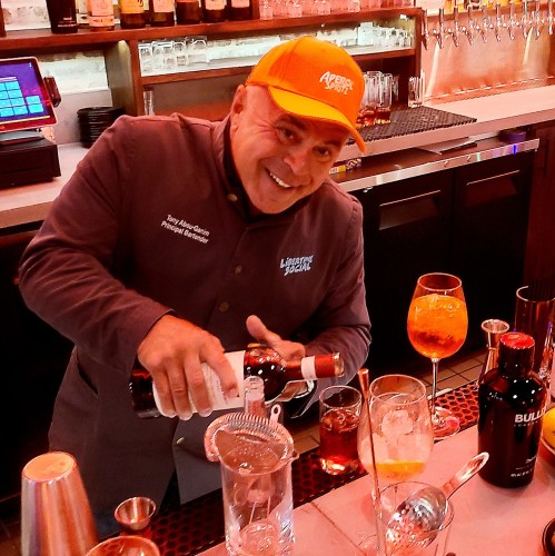 A smiling bartender pours a drink at a bar, surrounded by various bottles and glasses.
