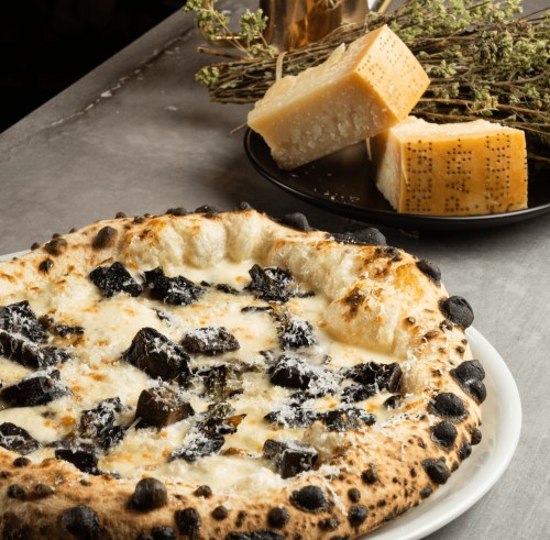 A close-up of a pizza topped with black ingredients, served with a plate of cheese and herbs in the background.