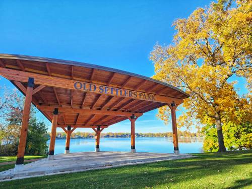 A wooden pavilion at Old Settlers Park, surrounded by colorful autumn trees and a serene lake under a clear blue sky.