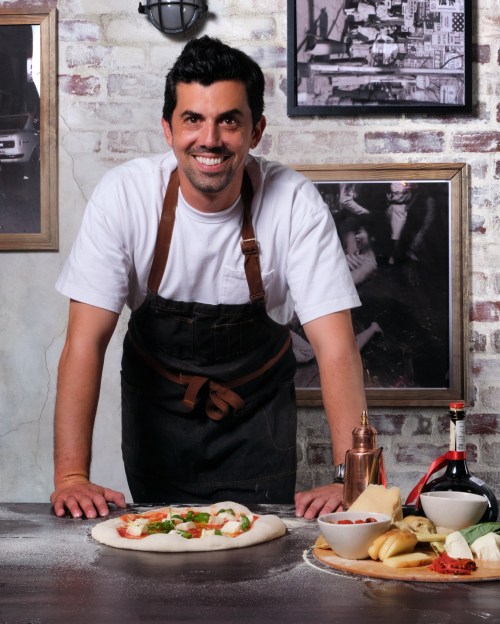 A smiling chef in an apron stands behind a table with a pizza and various ingredients, set against a rustic backdrop.