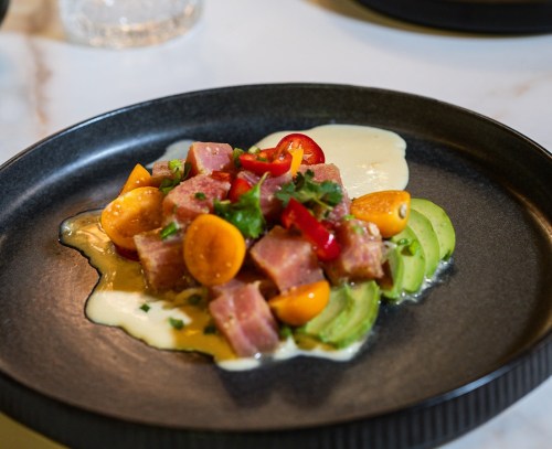 A black plate with diced ham, avocado, cherry tomatoes, and a creamy sauce, garnished with herbs and red pepper.