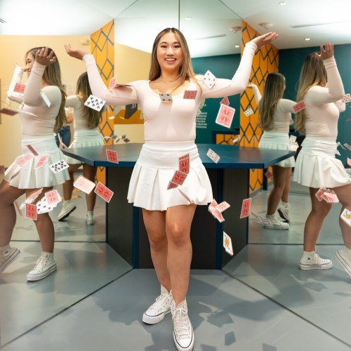 A smiling person in a white outfit stands in a mirrored room, surrounded by colorful playing cards.