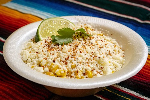 A bowl of corn topped with cheese, lime, cilantro, and spices, set on a colorful woven textile.