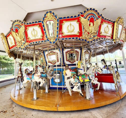 A colorful carousel with ornate decorations and various painted horses, set in a park-like area.