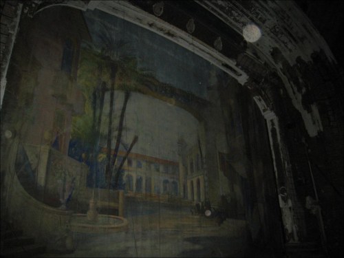 A faded mural depicting a tropical scene with palm trees and buildings, set in a dimly lit, abandoned theater.