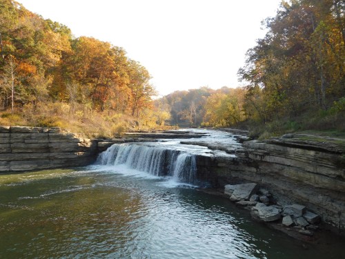 A serene waterfall cascades over rocky ledges, surrounded by autumn foliage and a calm river.