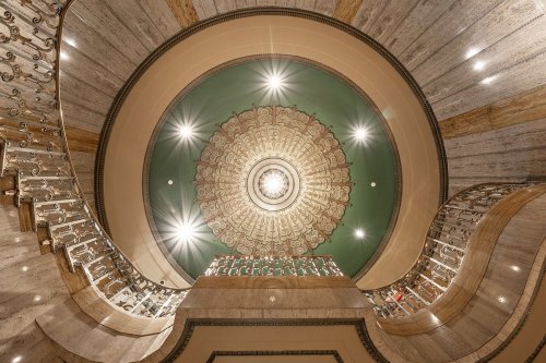 A grand staircase with ornate railings, viewed from below, featuring a decorative ceiling and bright lights.