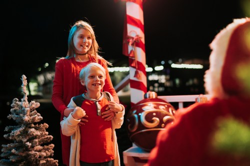 A girl in a red dress stands behind a smiling boy in a sweater, both looking at Santa in a festive setting.